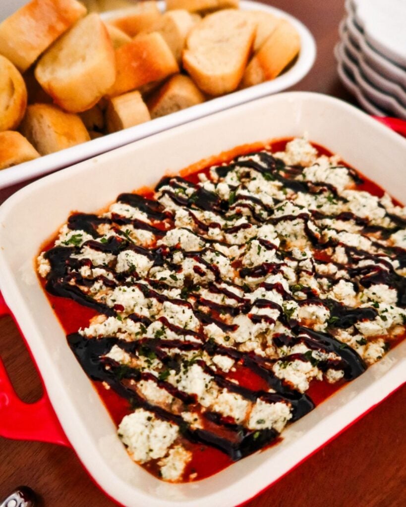 Baking dish of marinara dip topped with crumbled Boursin cheese and balsamic drizzle, served with toasted bread.