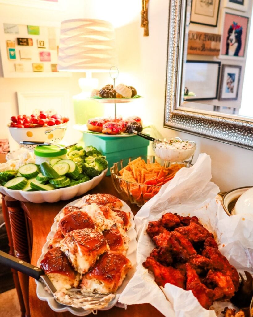 Buffet table with dips, sliders, wings, veggies, crackers, and desserts arranged for entertaining.