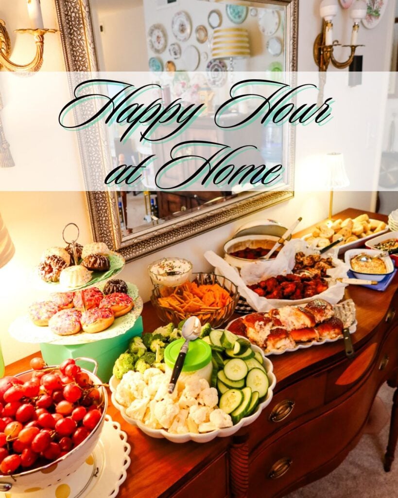 A cozy buffet table set for a happy hour at home, featuring appetizers like wings, sliders, dips, fresh vegetables, crackers, grapes, and a tiered stand of colorful mini cakes, styled with warm lighting and a vintage mirror backdrop.