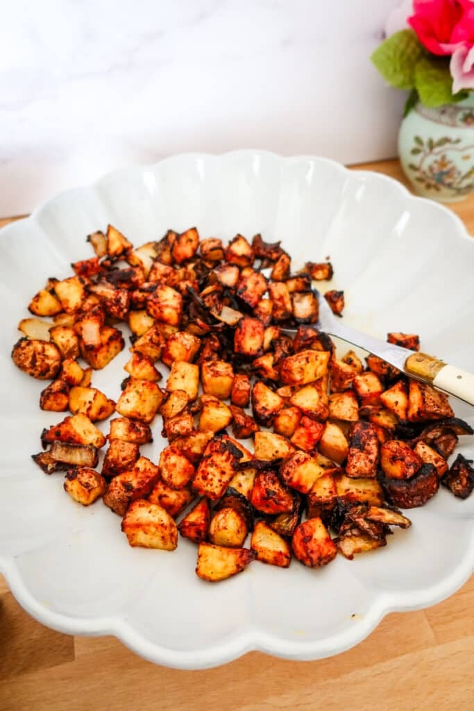 Golden brown air fryer potatoes and onions with a smoky red Firecracker seasoning, served in a white scalloped bowl on a wood counter.