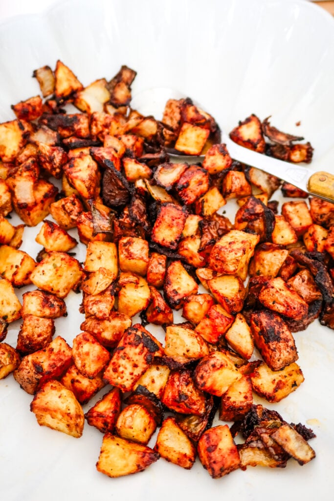 Golden brown air-fried potatoes and onions with spicy seasoning, served on a white scalloped plate with a vintage knife.