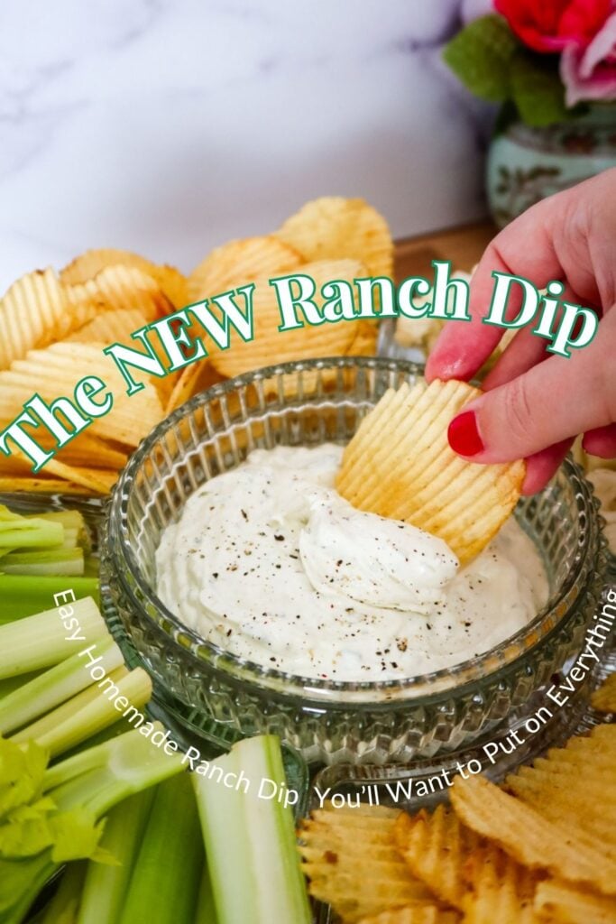 A hand dipping a ridged potato chip into a bowl of creamy homemade ranch dip surrounded by chips, celery, and flowers in the background.