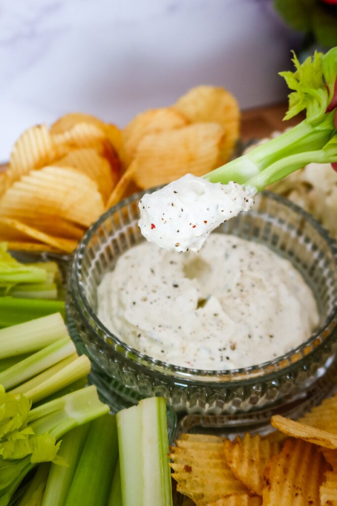 Celery stick dipped in thick homemade ranch dip with chips and cauliflower on the side.