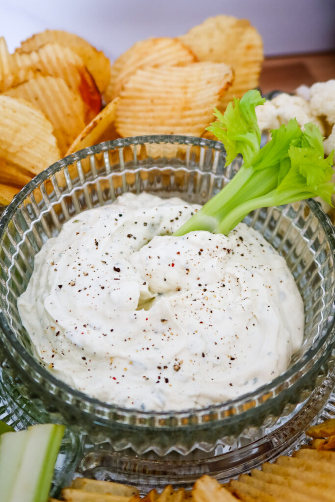 A celery stick with ranch dip in front of a bowl of creamy ranch surrounded by chips and vegetables.