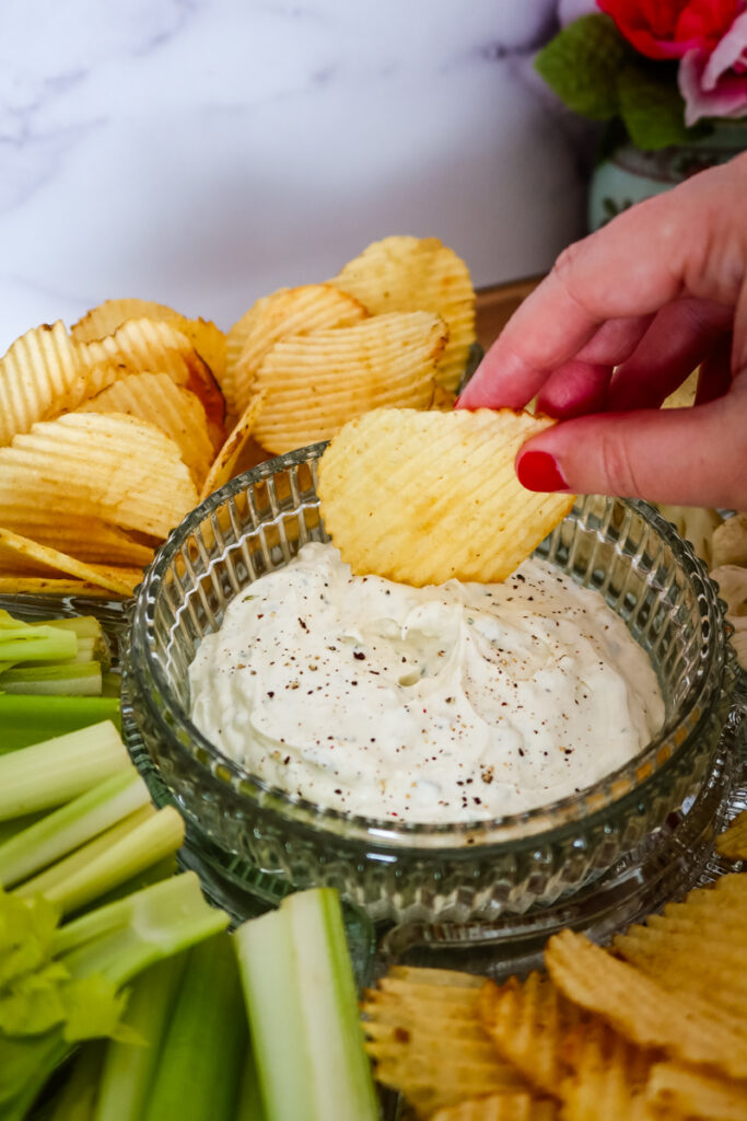A hand dipping a potato chip into a bowl of creamy homemade ranch dip surrounded by chips and veggies.