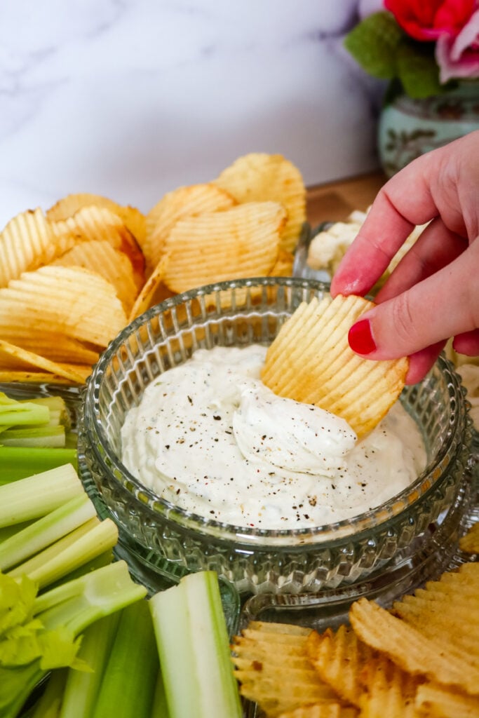 A ridged potato chip dipped into a bowl of creamy ranch dip surrounded by chips and celery sticks.