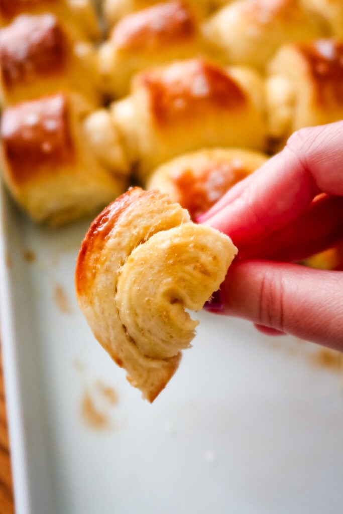 A hand holding a twisted, soft piece of Parkerhouse roll in front of the pan.
