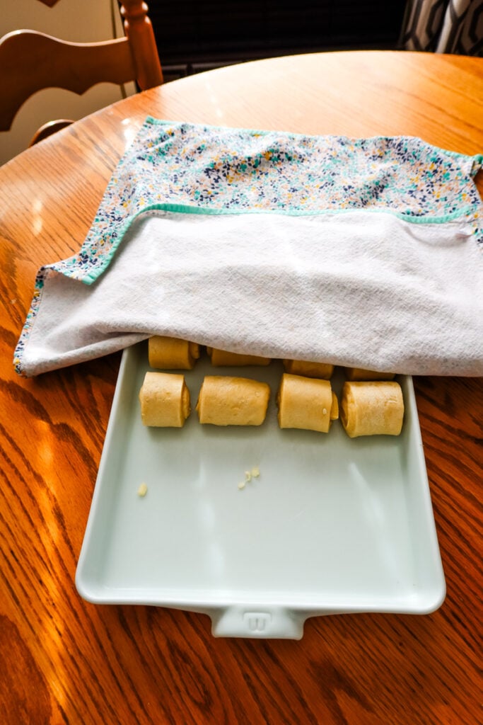 Tray of shaped Parkerhouse rolls partially covered with a kitchen towel while rising on a wooden table.