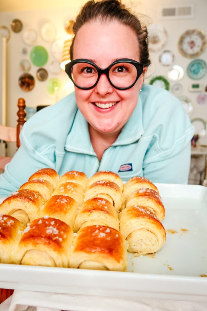 A smiling woman holding a fresh pan of golden Parkerhouse rolls in a bright kitchen.