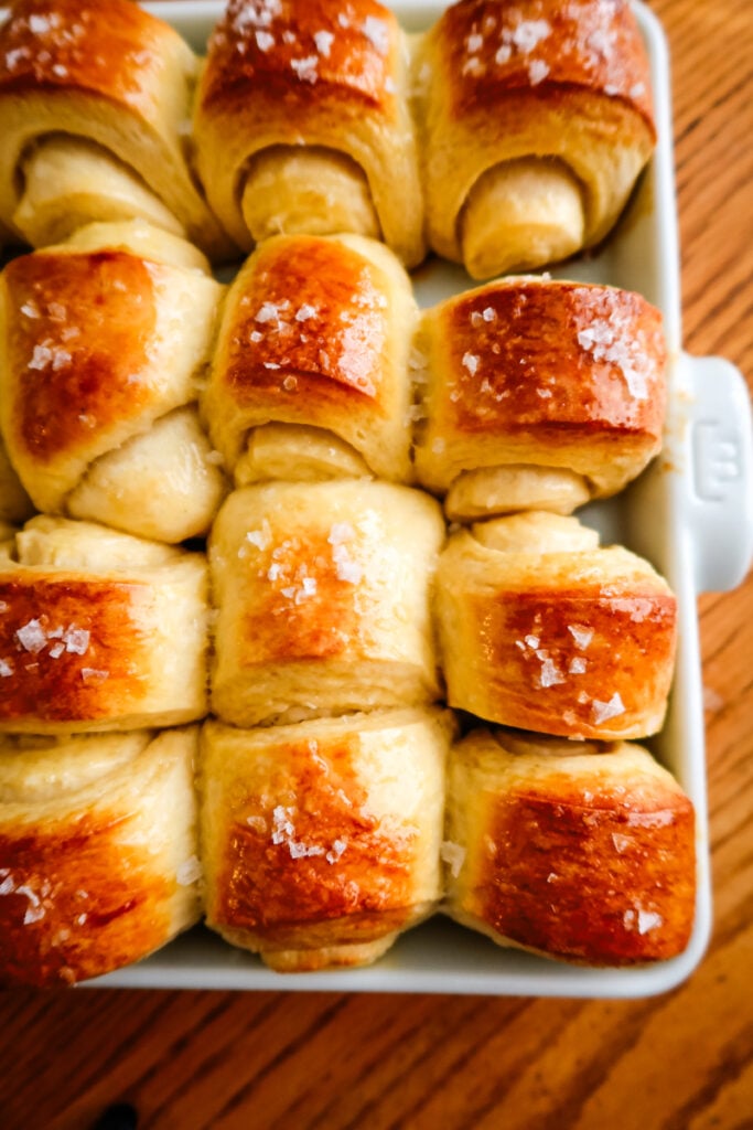 Golden brown Parkerhouse rolls in a baking dish, brushed with honey butter and sprinkled with flaky sea salt.