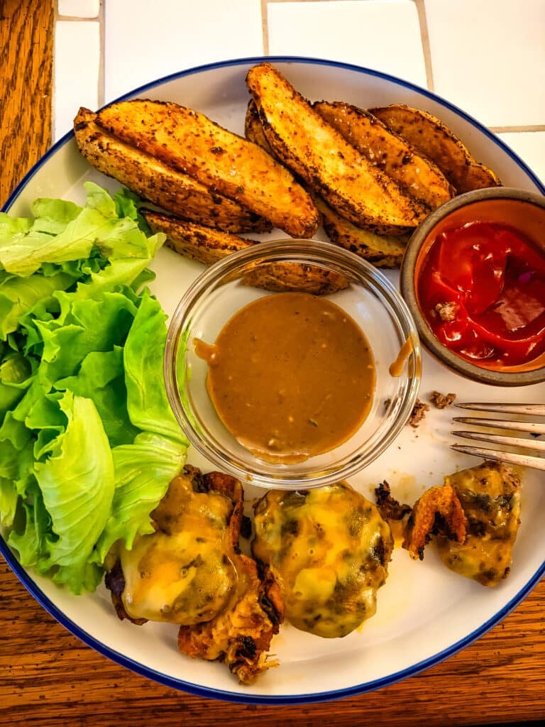 Mini cheeseburgers served with seasoned potato wedges, green lettuce, ketchup, and brown dipping sauce on a white dinner plate.