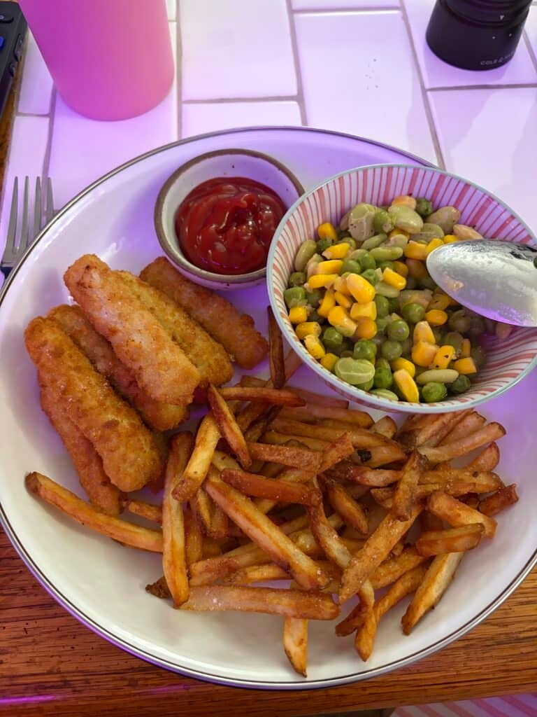 Crispy breaded fish sticks and French fries served with ketchup and a bowl of mixed corn, peas, and lima beans on a dinner plate.