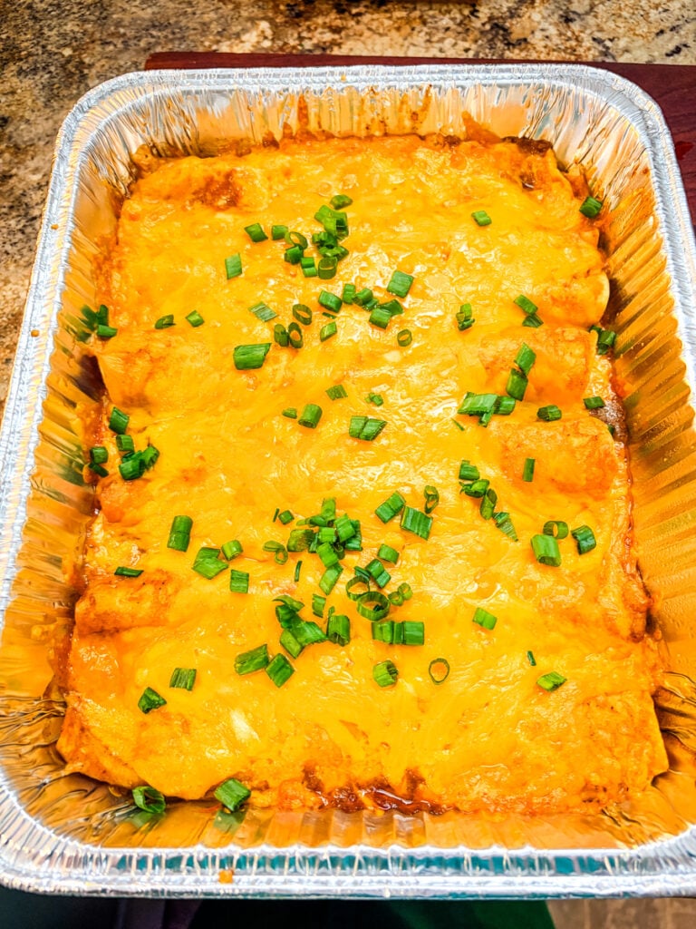 Overhead view of a foil baking pan filled with cooked pulled pork enchiladas, melted cheddar cheese, and red enchilada sauce.