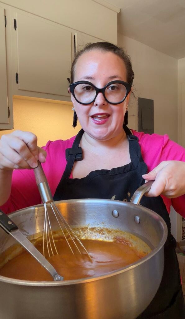 Ashley whisking homemade red enchilada sauce in a large stainless steel pot using Bolner's Fiesta Brand Enchilada Seasoning.