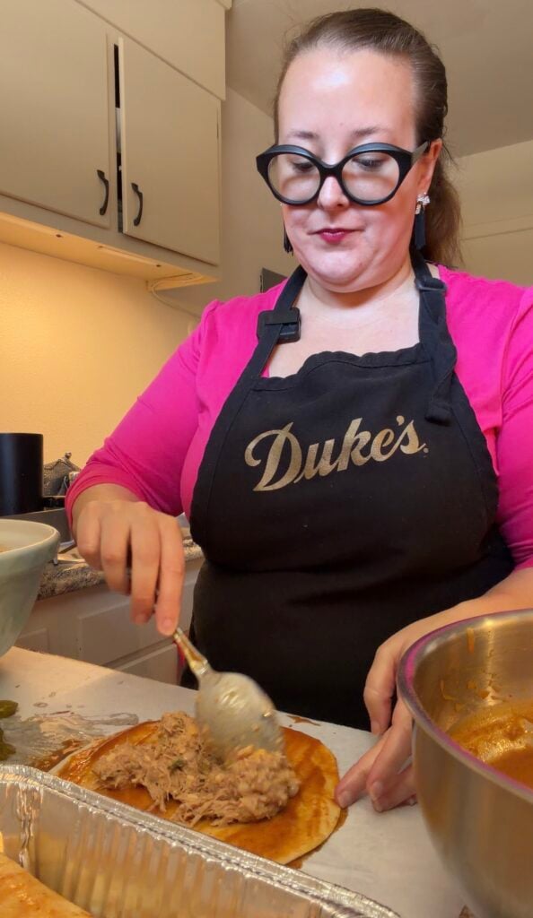 Ashley from Sweetpea Lifestyle spooning creamy pulled pork filling onto a flour tortilla coated in red enchilada sauce.
