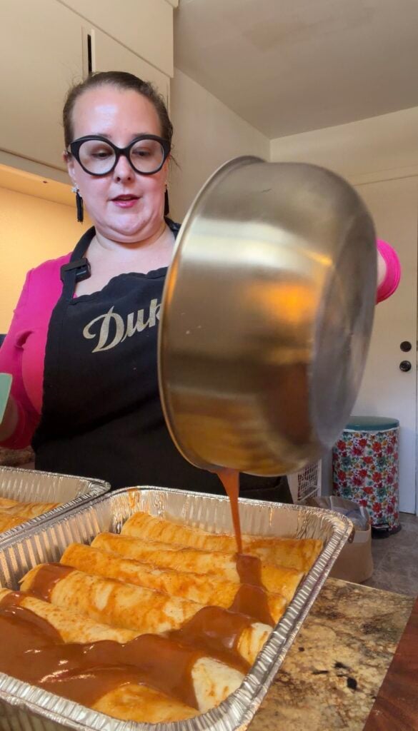 Ashley pouring homemade red enchilada sauce from a large stainless steel pot over six rolled pulled pork enchiladas in an aluminum baking pan.