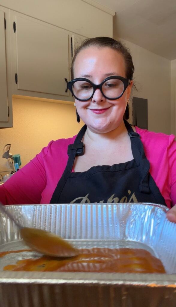 Ashley spreading red enchilada sauce across the bottom of a disposable aluminum baking pan with a ladle.