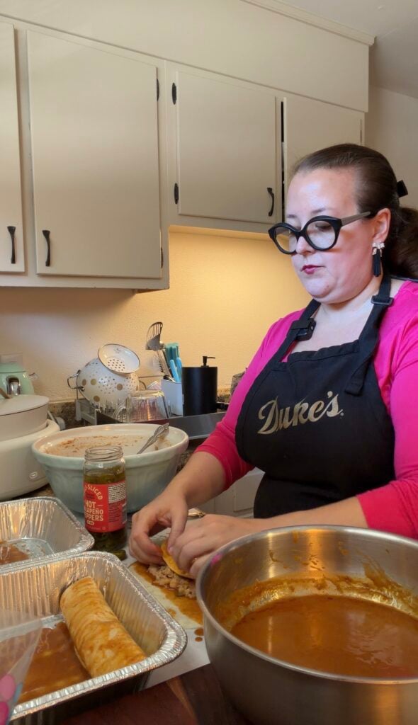 Ashley rolling a flour tortilla around creamy pulled pork filling, with a bowl of red enchilada sauce and aluminum baking pans on the counter.