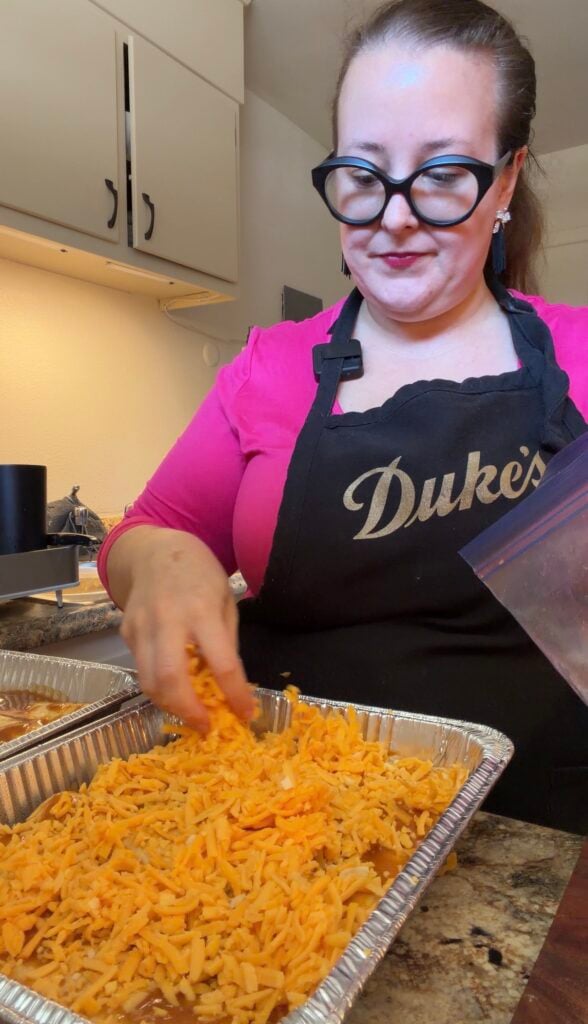 Ashley sprinkling freshly shredded orange cheddar cheese over a large aluminum pan of pulled pork enchiladas before baking.